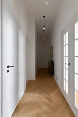 Korytarz z parkietem ułożonym w jodełkę francuską, białymi drzwiami i serią szklanych lamp wiszących.