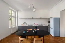 Kuchnia w kamienicy z grafitową zabudową, marmurowym blatem, pastelowo niebieską lodówką w stylu retro i czarnym owalnym stołem z drewnianymi krzesłami.