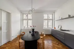 Jadalnia z czarnym owalnym stołem i drewnianymi krzesłami na parkiecie w jodełkę, z liniową zabudową kuchenną w grafitowym kolorze i wysokimi oknami.