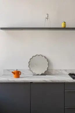 Detal kuchni z marmurowym blatem, pomarańczowym ceramicznym dzbankiem, falowanym lustrem oraz wyciskaczem do cytryn na grafitowej półce.