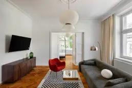 Salon z dekoracyjną rozetą sufitową, ciemną komodą pod telewizorem, szarą sofą, czerwonym fotelem RM58 i widokiem na sypialnię w tle.