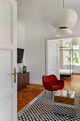 Fragment salonu z czerwonym fotelem RM58, lustrzanym stolikiem na dywanie w kratkę i ciemną komodą z dekoracyjną zieloną wazą.
