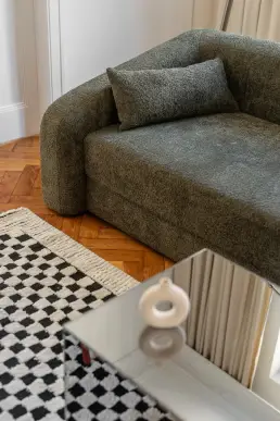 Zbliżenie na ciemnozieloną sofę bouclé z poduszką, fragment dywanu w czarno-białą kratkę i lustrzany stolik z białą ceramiczną wazą.