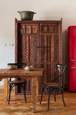 Bogato rzeźbiona drewniana szafa z geometrycznymi wzorami zestawiona z drewnianym stołem, gięciętymi krzesłami i czerwoną lodówką retro.