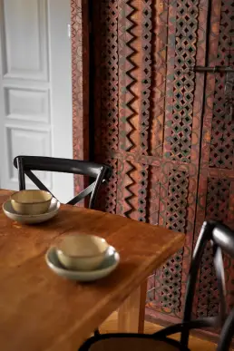 Detal zabytkowej szafy z ręcznie rzeźbionymi ornamentami zygzakowymi i rombowymi obok prostego drewnianego stołu z ceramicznymi miseczkami.