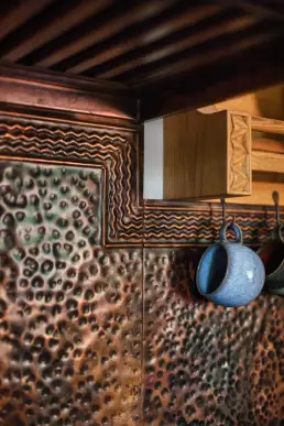 Zbliżenie na fartuch z kutej miedzi z wytłaczanym wzorem fal i punktów oraz ceramiczne kubki zawieszone na metalowych haczykach.