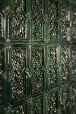 Detal zielonych kafli zdobiących piec kaflowy, z reliefowym ornamentem roślinnym i geometrycznymi obramowaniami.