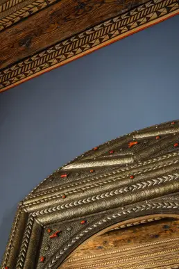 Zbliżenie na malowany drewniany sufit z trójkątnym ornamentem oraz górną część bogato zdobionego lustra w srebrnej ramie z koralowymi akcentami.