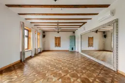 Przestronna sala z drewnianymi belkami stropowymi, dekoracyjnym parkietem w romby, miętowym piecem kaflowym i dużym lustrem w zdobionej ramie.