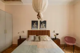 Sypialnia z karmelowym tapicerowanym zagłówkiem, dwoma drewnianymi stolikami nocnymi w stylu vintage, fotograficznymi grafikami nad łóżkiem i szarą zabudową szafy z mosiężnymi uchwytami.