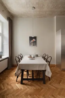 Jadalnia z drewnianym stołem nakrytym lnianym obrusem, giętymi krzesłami Thonet i dekoracyjnym, odsłoniętym sufitem z oryginalnym wzorem florystycznym.
