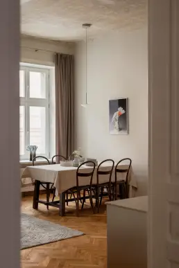 Widok przez przeszklone drzwi na jadalnię z lnianym obrusem, giętymi krzesłami Thonet i charakterystycznym, oryginalnym sufitem kamienicy.