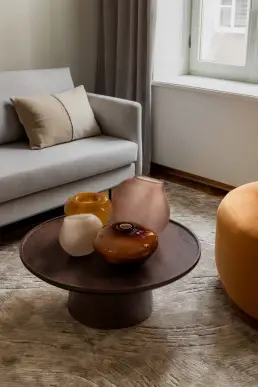 Detal salonu z drewnianym okrągłym stolikiem kawowym z kompozycją bursztynowych i pudroworóżowych szklanych waz oraz fragmentem aksamitnego pufa w kolorze koniaku.