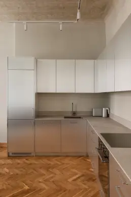 Nowoczesna kuchnia ze stalową zabudową, jasnymi szafkami górnymi, kamiennym blatem i odsłoniętym fragmentem dekoracyjnego sufitu nad parkietem w jodełkę.