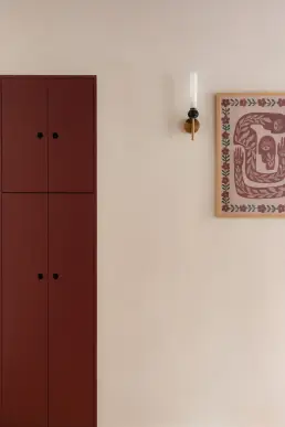 Bordowa wysoka szafa z czarnymi okrągłymi uchwytami, mosiężny kinkiet ze świecą i oprawiony plakat z ornamentem roślinnym na beżowej ścianie