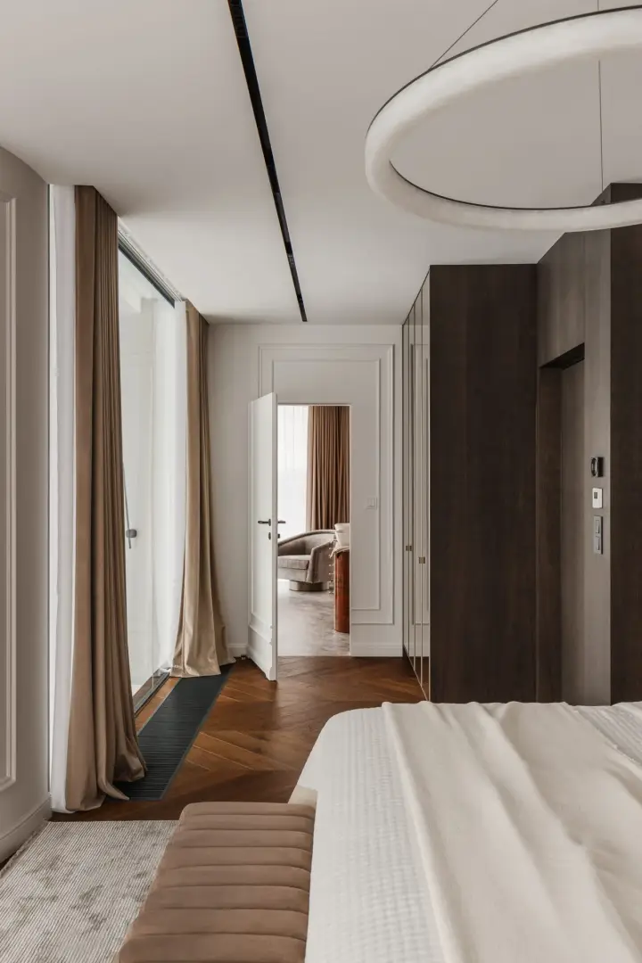 Sypialnia z zabudową szaf, okrągłym żyrandolem i przejściem do kolejnego pokoju – wnętrze apartamentu