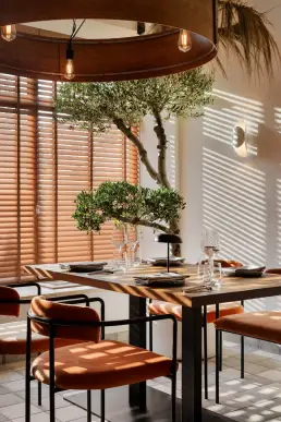 restauracja Muro Rzeszów — stolik z pomarańczowymi krzesłami przy drzewku oliwnym w naturalnym świetle żaluzji, okrągła lampa nad głową