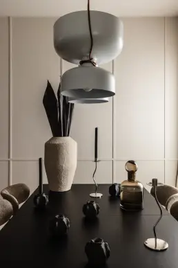 Detal jadalni z czarnym stołem, ceramicznymi czarnymi wazonami, szklaną karafką w bursztynowym odcieniu i designerską lampą wiszącą w neutralnym kolorze.