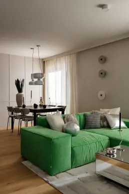 Strefa dzienna z zieloną sofą modułową na jasnym dywanie, lustrzanym stolikiem kawowym i jadalnią z czarnym stołem w tle.