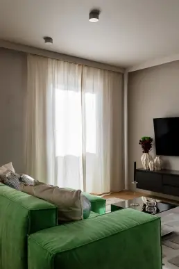 Salon z zieloną welurową sofą, lnianymi zasłonami w pełnej długości i czarną szafką RTV z kolekcją ceramicznych wazonów.
