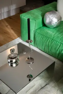 Lustrzany stolik kawowy z chromową rzeźbą oraz świecznikiem na tle zielonej welurowej sofy z metalicznymi poduszkami.